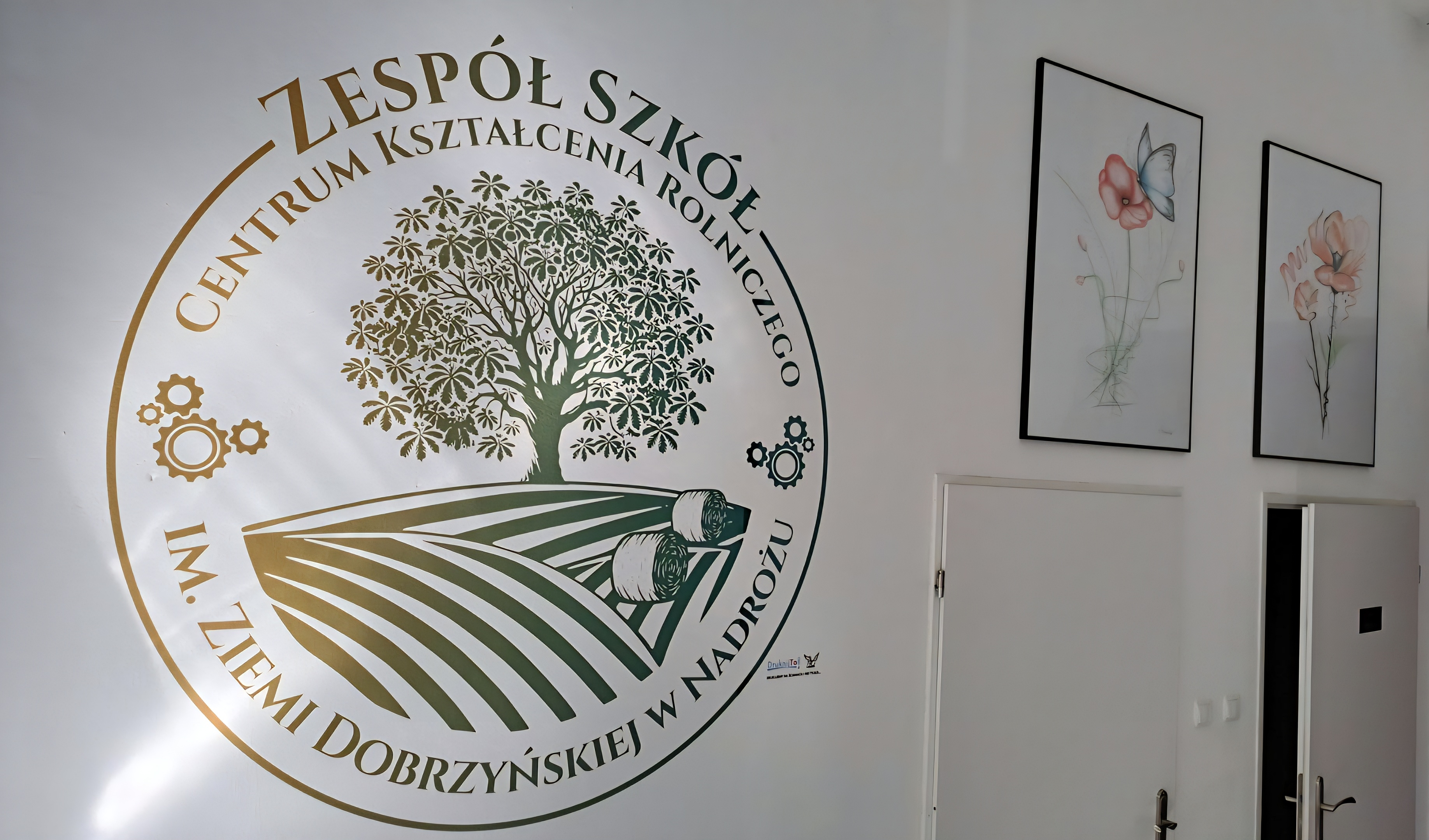 Logo Zespołu Szkół w Nadrożu wydrukowane złotym tuszem na białej ścianie